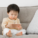 Young toddler holding and viewing a smart phone while sitting on the couch